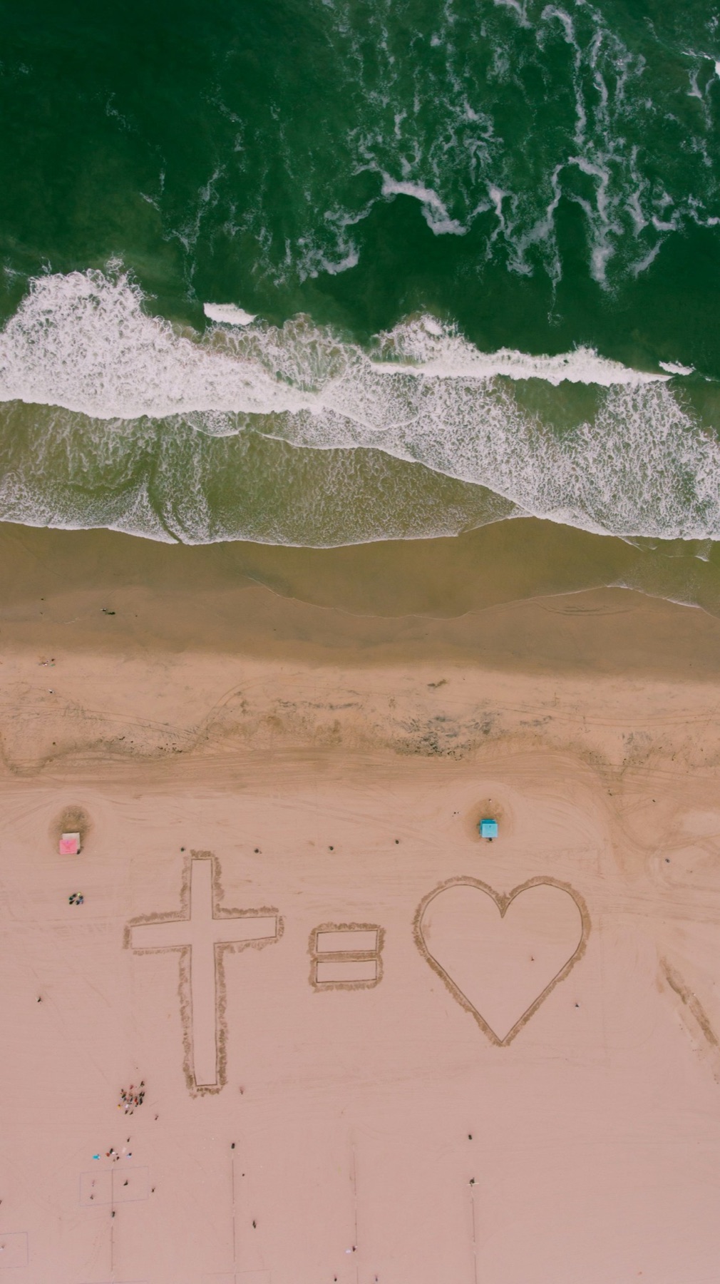 Aerial view of a beach with a cross, an equals sign and a heart drawn in the sand: the cross equals love